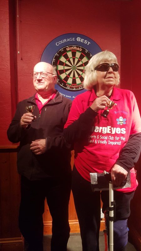 DAVID AND ANNIE STOOD IN FRONT OF THE DART BOARD HOLDING THEIR TROPHIES